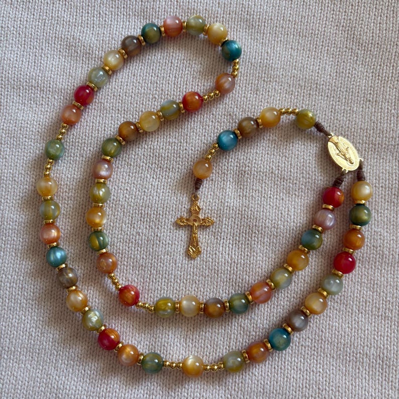 May include: A rosary featuring multicolored, translucent, polished stone beads in shades of red, blue, green, and yellow, separated by small gold-toned metal spacers. The piece includes a gold-toned metal centerpiece and a detailed gold-toned crucifix.