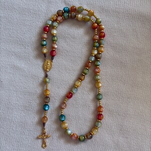 May include: A rosary featuring multicolored, translucent beads in shades of blue, red, green, yellow, and brown, separated by small gold-toned metal spacers. It includes a gold-toned metal centerpiece depicting a figure and a gold-toned crucifix.