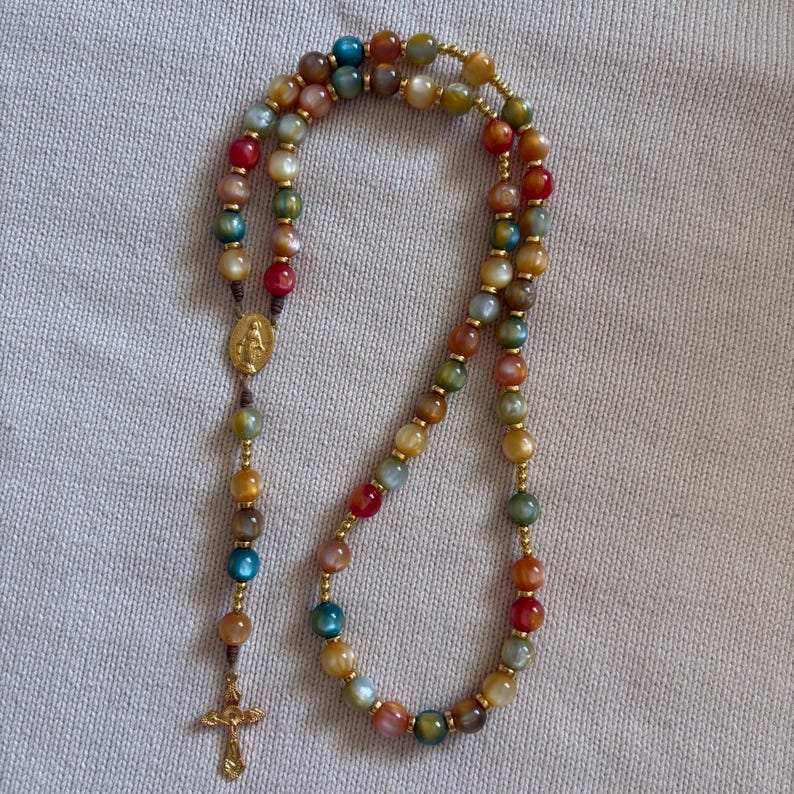 May include: A rosary featuring multicolored, translucent beads in shades of blue, red, green, yellow, and brown, separated by small gold-toned metal spacers. It includes a gold-toned metal centerpiece depicting a figure and a gold-toned crucifix.
