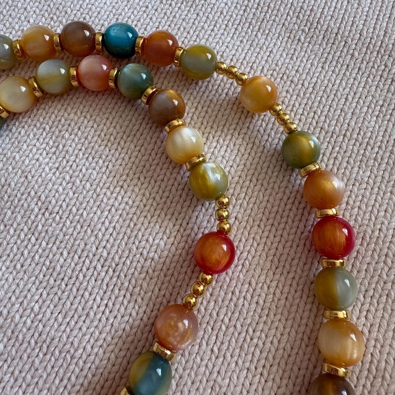 May include: A beaded necklace featuring smooth, multicolored round stones in shades of amber, teal, brown, and cream. The stones are interspersed with small, polished gold-toned metal spacer beads, creating a repeating pattern along the strand.