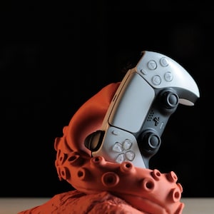 May include: A white and black video game controller is held by an orange octopus-shaped stand. The controller has multiple buttons and directional pads. The stand is on a light brown surface with a black background.