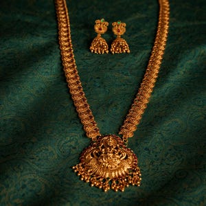 Könnte beinhalten: Goldfarbenes Halsketten- und Ohrring-Set. Die Halskette hat einen detaillierten Anhänger mit aufwendigen Designs und eine lange Kette. Die Ohrringe sind klein, rund und weisen ebenfalls detaillierte Designs auf. Der Schmuck wird auf grünem Stoff präsentiert.