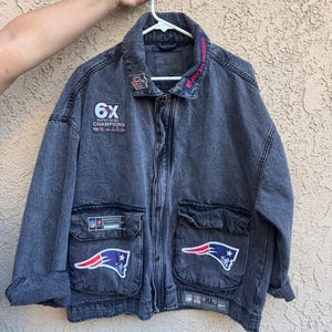 Könnte beinhalten: Graue Jeansjacke mit mehreren Aufnähern, darunter das Logo der New England Patriots und "6X Super Bowl Champions". Die Jacke hat zwei Vordertaschen und einen Knopfverschluss. Der Kragen lautet "WE ARE ALL PATRIOTS."