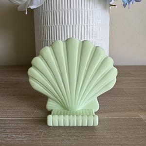 May include: A pale green seashell ornament. The shell has a fan-like design with vertical ridges and a rectangular base. The ornament is displayed on a wooden surface, with a white vase and flowers in the background.