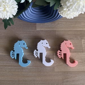 May include: Three seahorse-shaped decorations in light blue, white, and coral pink. The seahorses are on a wooden surface. White flowers and a blue vase are in the background.