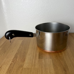 May include: A stainless steel saucepan with a copper-colored base and a black handle. The handle is attached to the side of the pan with two silver-colored metal rivets and features a small metal loop at the end for hanging the cookware.