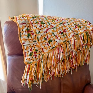 May include: A handmade crochet blanket featuring a granny square pattern in cream, orange, red, and green yarn. The blanket is finished with long, thick fringe tassels along the edge, incorporating the same color palette of orange, red, white, and green.