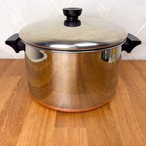May include: Stainless steel cooking pot with a lid and black handles. The pot has a copper-colored base and a black knob on the lid. The pot is sitting on a wooden surface.