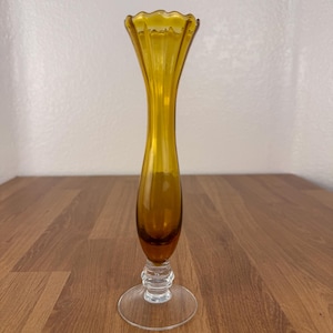 May include: Amber-colored glass vase with a flared, scalloped rim. The vase has a slender, elongated body and sits on a clear, circular base. The vase is approximately 10 inches tall, perfect for displaying flowers or as a decorative piece.