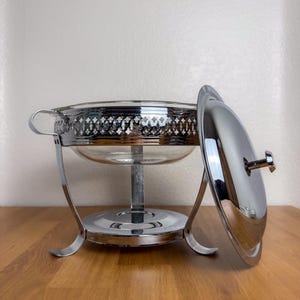 May include: A silver-colored chafing dish with a clear glass bowl and a matching lid. The dish has a decorative, pierced metal rim and is supported by a stand with curved legs. The lid is partially open, revealing the interior.