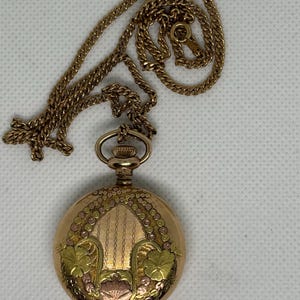 Könnte beinhalten: Eine goldfarbene Taschenuhr mit einem verzierten Design, die an einer passenden Kette hängt. Das Zifferblatt der Uhr zeigt florale und geometrische Muster in Gold-, Rosa- und Grüntönen. Die Kette ist ein filigraner, gegliederter Stil.