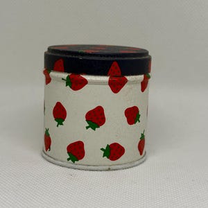Può includere: Piccolo contenitore rotondo in latta con sfondo bianco e motivo a fragole rosse. Il coperchio è blu scuro con disegni di fragole rosse. Il contenitore ha un aspetto vintage, leggermente screpolato.