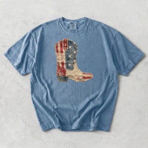 May include: A light blue short-sleeve t-shirt featuring a central graphic of a cowboy boot. The boot design is composed of patchwork fabric elements, including a blue section with white stars, red and white stripes, and lace detailing.