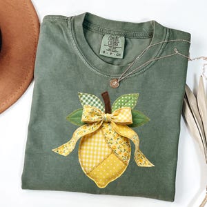 May include: Olive green Comfort Colors t-shirt with a lemon applique. The lemon is made of yellow fabric with a gingham and floral pattern, and a brown stem. A silver necklace with a small pendant is on the shirt.