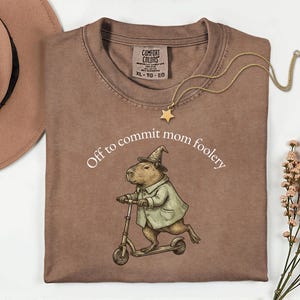 May include: Brown t-shirt with the text "Off to commit mom foolery" and a capybara riding a scooter, wearing a witch hat and coat. A gold star pendant necklace is draped across the shirt. A brown hat is in the upper left corner.