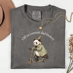 May include: A charcoal gray t-shirt with the phrase "Off to commit dad foolery" printed above an illustration of a panda riding a scooter. The panda wears a wizard hat and coat. A gold star pendant necklace rests on the shirt.