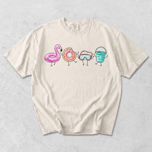 May include: A cream-coloured t-shirt with a cartoon illustration of a pink flamingo float, a donut, swim goggles, and a blue bucket, each with smiling faces. The design is in a cartoon style, ideal for summer or beach wear.
