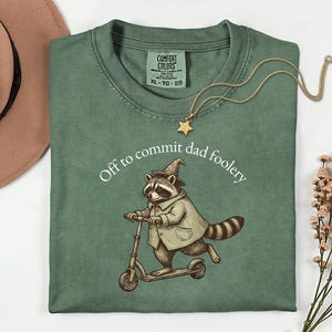 May include: Sage green t-shirt with a graphic of a raccoon riding a scooter, wearing a wizard hat and coat. The text "Off to commit dad foolery" is above the raccoon. A gold star pendant necklace is draped on the shirt.