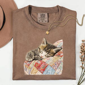 May include: A brown Comfort Colors t-shirt featuring a sleeping kitten on a patchwork quilt. A gold star pendant necklace rests on the shirt. A brown hat is in the upper left corner. The shirt has a tag that says XL-TG-EG.