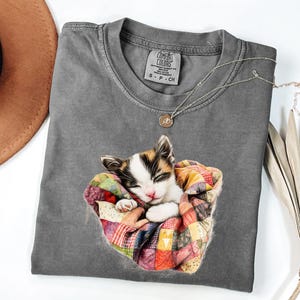 May include: A gray Comfort Colors t-shirt featuring a sleeping kitten nestled in a colorful patchwork quilt. A silver necklace with a small pendant rests on the shirt. The shirt is a size small.