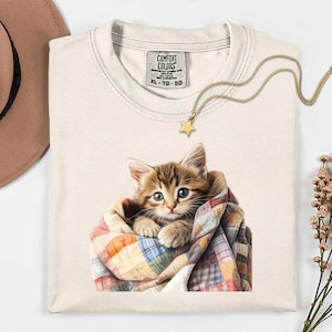 May include: Cream-colored Comfort Colors XL t-shirt featuring a watercolor illustration of a kitten wrapped in a colorful patchwork quilt. A gold star pendant necklace and a brown hat are also visible.