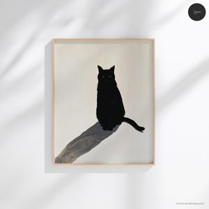 Op de afbeelding: Een ingelijste kunstdruk met een effen zwarte kat die rechtop zit tegen een lichtbeige achtergrond. De kat werpt een lange, donkere, diagonale schaduw over het oppervlak. Het kunstwerk is gevat in een eenvoudige, lichtgekleurde houten lijst.