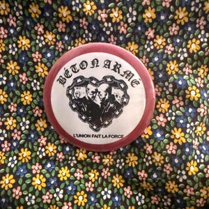 May include: A round, maroon button with the text "BETON ARME" in a gothic font. The center features a black and white image of three figures within a chain-linked heart, with the text "L'UNION FAIT LA FORCE" below.