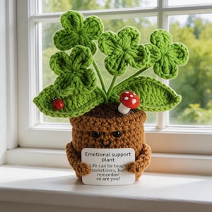 May include: Crocheted emotional support plant in a brown pot with green leaves and a red mushroom. A small white sign reads "Emotional support plant. Life can be tough sometimes, but remember so are you!" A red ladybug is on a leaf.
