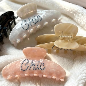 Könnte beinhalten: Eine Sammlung von Haarspangen in verschiedenen Farben, darunter Rosa, Creme und Beige. Die Spangen haben einen Marmoreffekt und sind mit dem Wort "Chic" in kleinen, blauen Strasssteinen verziert. Die Spangen sind zum Halten der Haare konzipiert.