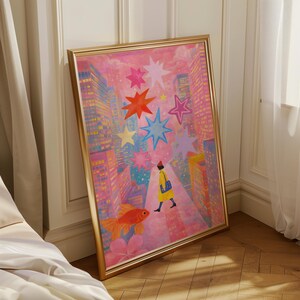 May include: A framed art print depicts a whimsical cityscape with a person in a yellow coat walking towards a goldfish. Colorful stars and buildings fill the pink background. The artwork is in a gold frame.