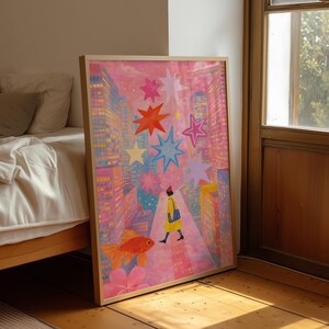 May include: A framed art print featuring a whimsical cityscape scene. The artwork depicts a person in a yellow coat walking down a street, with a goldfish and colorful stars. The buildings are pink and yellow, creating a vibrant and dreamlike atmosphere.