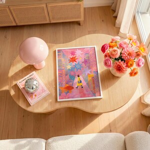 May include: A light wood, wavy-edged coffee table with a pink lamp, a book titled "Flower Market", and a framed art print. The print features a person walking on a street with a goldfish and colorful stars. A vase of flowers sits nearby.