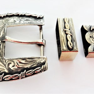 May include: Silver belt buckle with a barbed wire design and engraved floral patterns. The buckle is made up of three pieces: a main buckle, a keeper, and a tip.