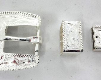 May include: Silver belt buckle set with a decorative engraved design. The set includes a buckle, a strap end, and a tip.