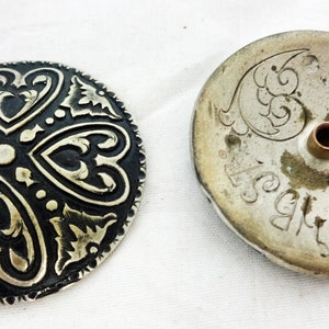 Könnte beinhalten: Zwei runde Metallknöpfe mit komplizierten Designs. Ein Knopf ist schwarz mit silbernen Akzenten und der andere ist silber mit einem Loch in der Mitte.