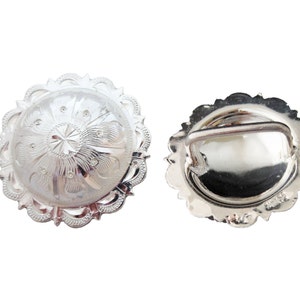 May include: Silver-toned metal buckle with a decorative design. The buckle has a round shape with a raised center and a scalloped edge. The back of the buckle has a smooth surface with a small metal loop.