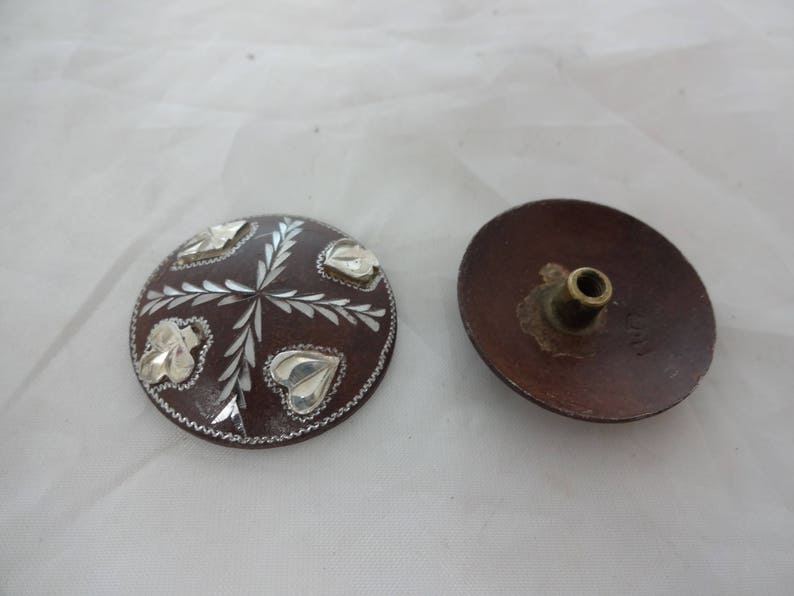 May include: Two round, brown, wooden buttons with silver leaf designs. One button has a small, brass, round knob on the back.