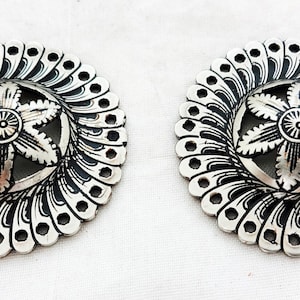 Könnte beinhalten: Zwei silberfarbene Metallknöpfe mit einem floralen Design. Die Knöpfe haben eine runde Form mit einer erhabenen Mitte, die eine fünfzackige Blume mit einem kleineren Kreis in der Mitte zeigt. Der äußere Rand der Knöpfe ist mit einem sich wiederholenden Muster aus kleinen Kreisen verziert.