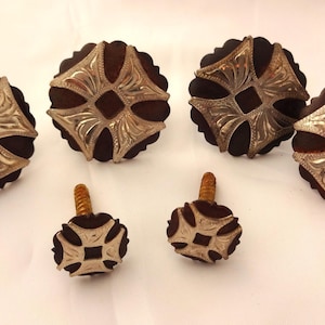 Pode incluir: Um conjunto de seis conchos e parafusos de metal prateado. Os conchos têm um fundo castanho escuro e um design decorativo, semelhante a uma flor. Os parafusos têm um design semelhante e um parafuso dourado.