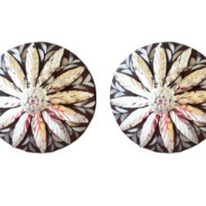 Puede incluir: Un par de pendientes redondos de plata con un diseño floral blanco y dorado sobre un fondo marrón.