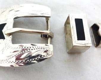 May include: Silver belt buckle with a black star design. The buckle is engraved with a floral pattern. The buckle is part of a set that includes two additional pieces.