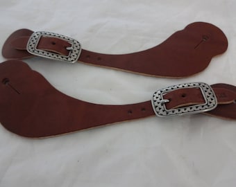 Può includere: Un paio di cinturini per speroni in pelle marrone con fibbie argentate. Le fibbie hanno un design geometrico.