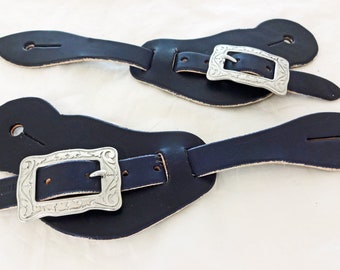 Può includere: Un paio di cinturini per speroni in pelle nera con fibbie argentate. Le fibbie hanno un design decorato.