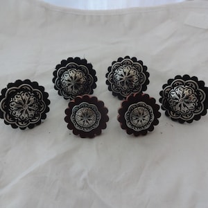 Puede incluir: Seis conchos met&aacute;licos decorativos con dise&ntilde;os florales. Tres conchos est&aacute;n sobre cuero negro y tres sobre cuero marr&oacute;n.