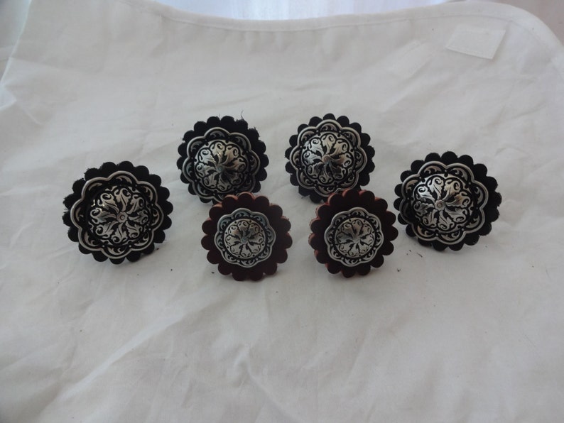 Puede incluir: Seis conchos met&aacute;licos decorativos con dise&ntilde;os florales. Tres conchos est&aacute;n sobre cuero negro y tres sobre cuero marr&oacute;n.