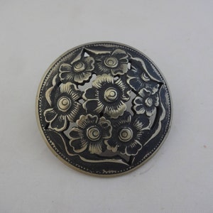 Könnte beinhalten: Eine runde, silberfarbene Brosche mit einem floralen Design. Die Blumen sind ausgeschnitten und haben einen schwarzen Hintergrund.
