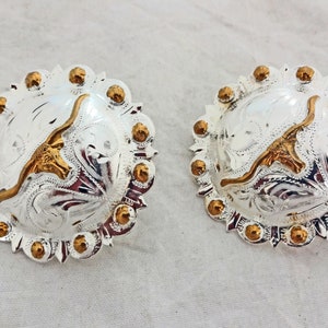 Könnte beinhalten: Ein Paar Cowboy-Gürtelschnallen aus Silber und Gold mit einem Longhorn-Stier-Design. Die Schnallen sind rund und haben ein erhabenes Design.