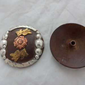 May include: A pair of metal belt buckles. One buckle is oval shaped with a floral design in copper, silver, and gold. The other buckle is round and has a hole in the center.