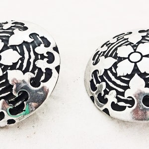 May include: A pair of silver tone earrings with a black and white floral design. The earrings are round and have a raised design.