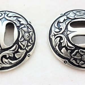 May include: Two silver-toned metal buckles with a floral design and a double-slot opening. The buckles are round and have a black background.